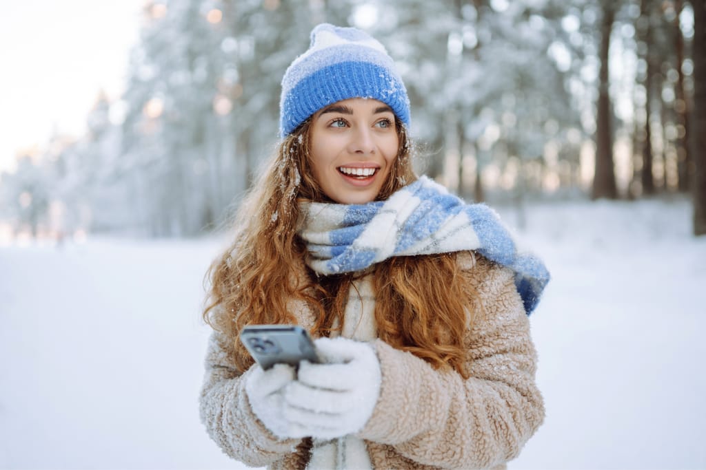 Hymyilevä nainen seisoo lumisessa metsässä sininen pipo ja huivi yllään, pitää käsissään älypuhelinta ja katsoo sivulle talvisessa maisemassa.
