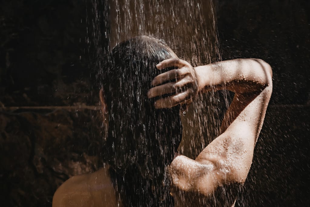Henkilö seisoo suihkussa hämärässä valaistuksessa, vesi valuu hiusten ja käsivarren yli.