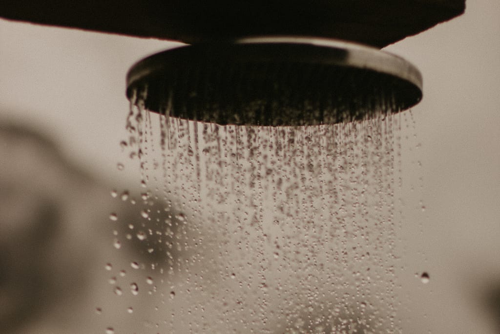 Lähikuva suihkupäästä, josta vesi virtaa tasaisena suihkuna vaaleaa taustaa vasten.