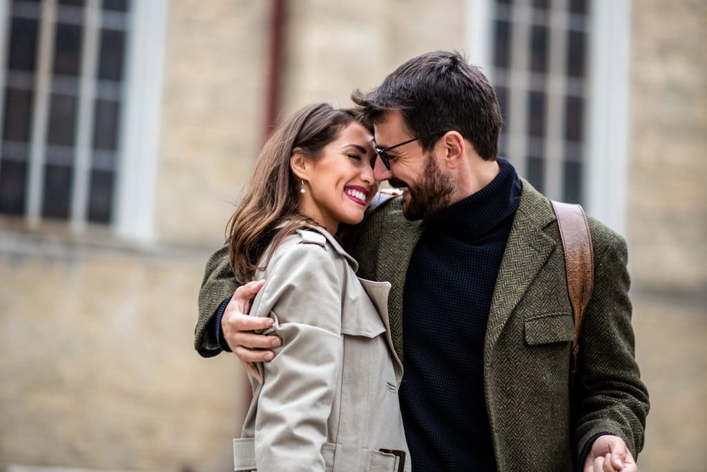 Nainen ja mies halaavat ulkona ja hymyilevät lähekkäin, taustalla näkyy rakennuksen ikkunoita.
