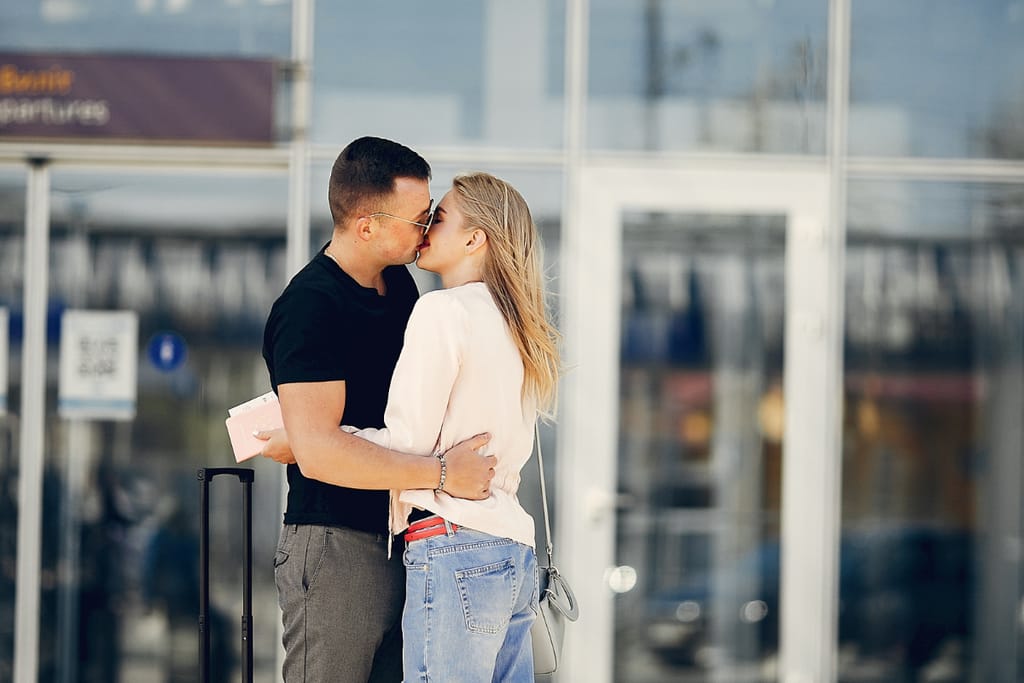 Pariskunta suutelee lentokentällä lähtöporttien edessä, toisella kädessä passi ja matkalaukku.