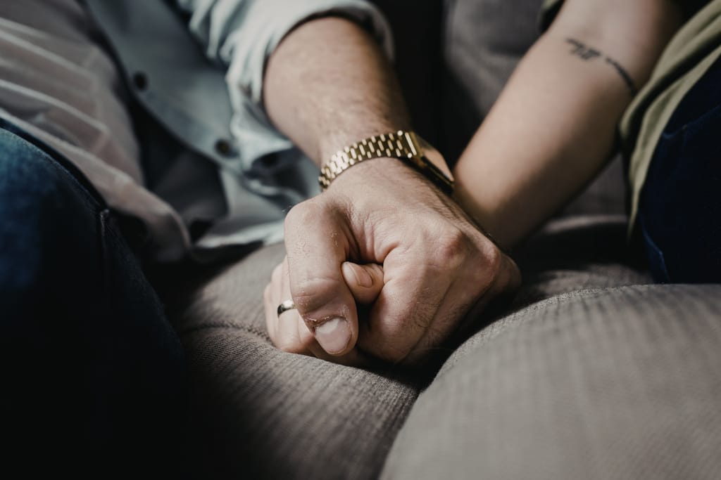 Lähikuva kahden henkilön käsistä, jotka pitävät toisiaan tiukasti kädestä sohvalla istuessaan.