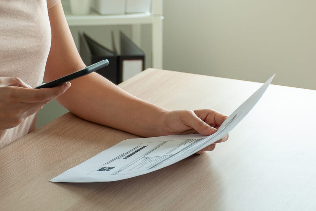 Henkilö pitelee kädessään laskupaperia ja tarkistaa tietoja älypuhelimesta pöydän ääressä.