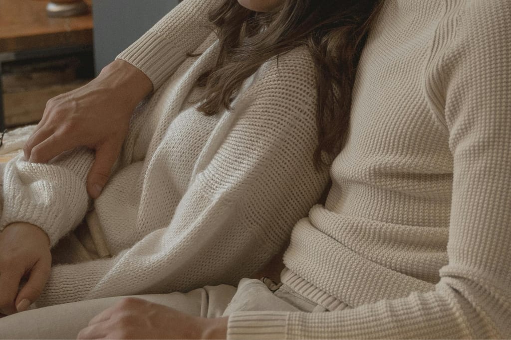 Pariskunta istuu lähekkäin sohvalla, toinen pitää kättään toisen sylissä rennossa ja läheisessä tunnelmassa.