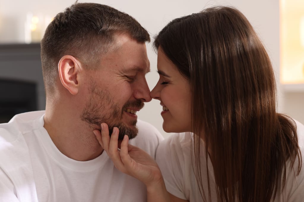 Kaksi aikuista istuu lähekkäin ja koskettaa toistensa kasvoja, he hymyilevät ja nojaavat toisiaan kohti.