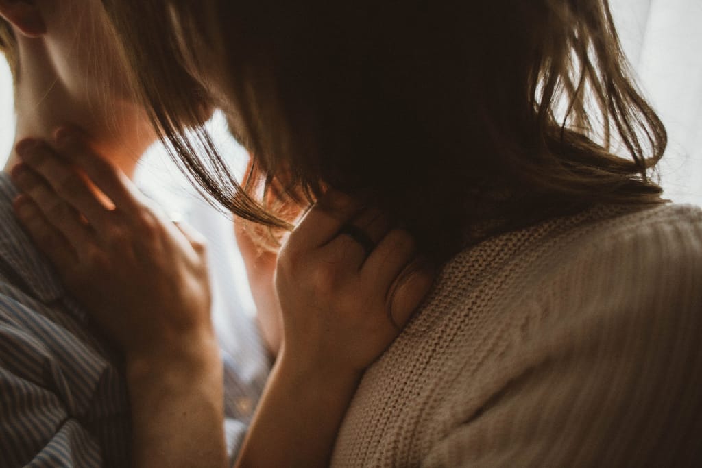 Kaksi aikuista suutelee, kädet toistensa niskalla ja hartioilla, kuva rajattu ylävartaloon.