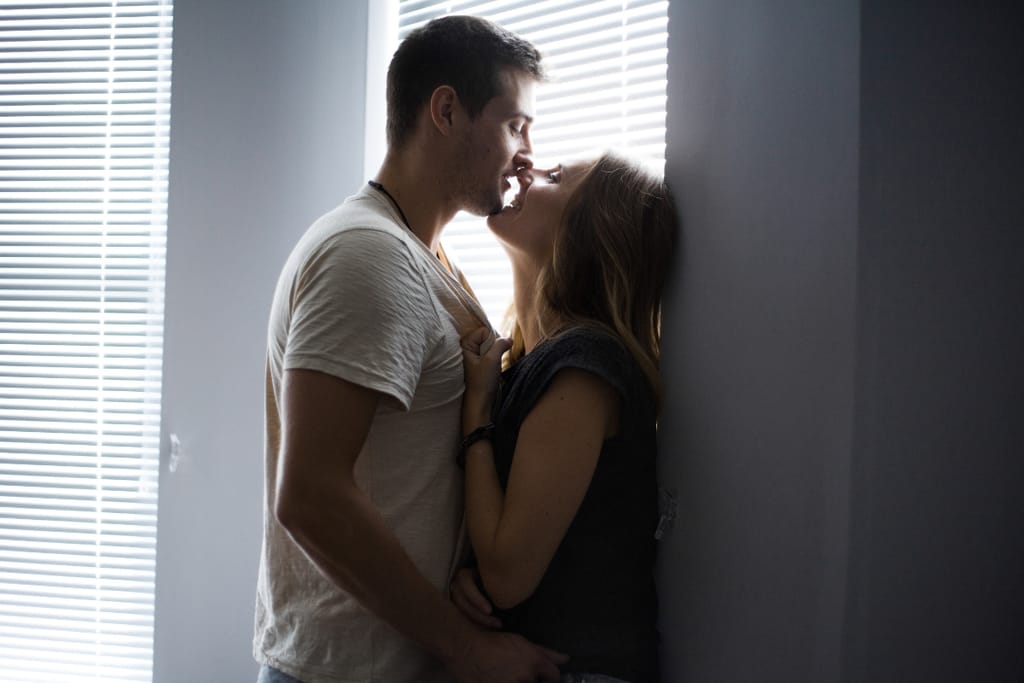 Nuori pari suutelee toisiaan intiimisti seinää vasten ikkunan sälekaihtimien suodattamassa valossa; mies pitää kättään naisen vyötäröllä ja nainen tarttuu miehen paitaan.