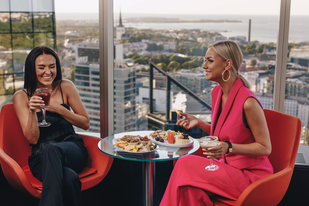 Alt-teksti:
Kaksi naista istuu pöydän ääressä korkealla ravintolassa, juo cocktaileja ja syö pieniä annoksia suurten ikkunoiden edessä kaupunkimaiseman näkyessä taustalla.