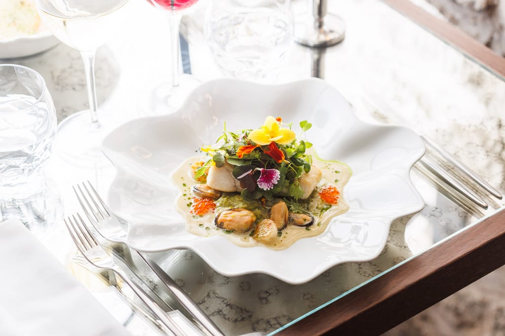 Taiteellisesti aseteltu ravintola-annos valkoisella aaltoilevareunaisella lautasella, annoksessa kalaa, simpukoita, kastiketta, yrttejä ja syötäviä kukkia, pöydällä viinilaseja ja ruokailuvälineet.