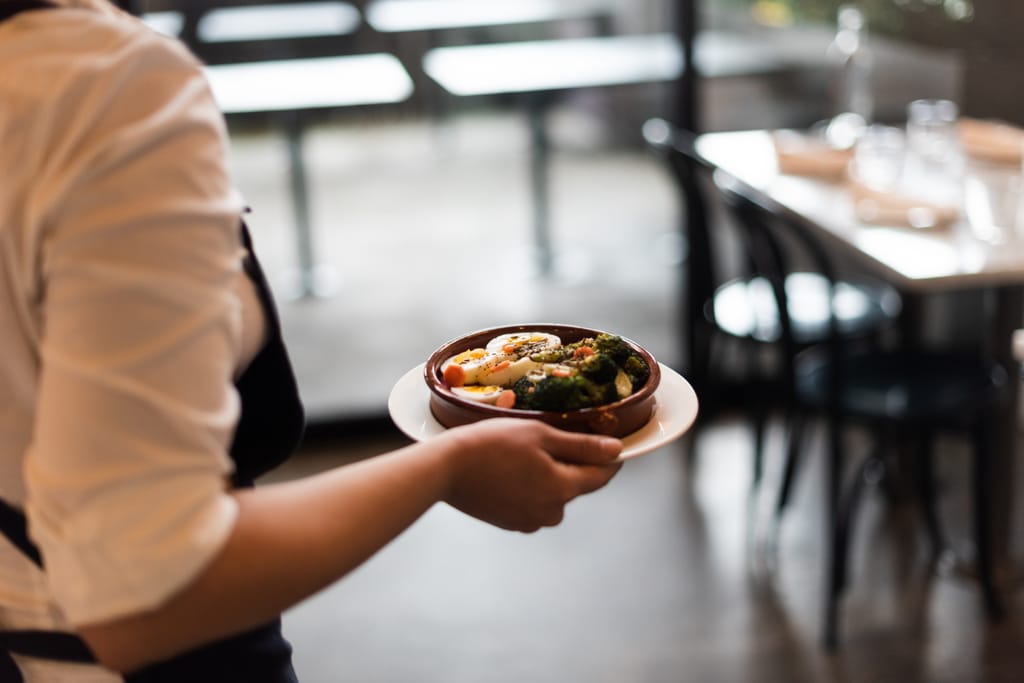 Tarjoilija kantaa käsissään annosta ravintolasalissa, taustalla pöytiä ja tuoleja pehmeästi epätarkkoina.