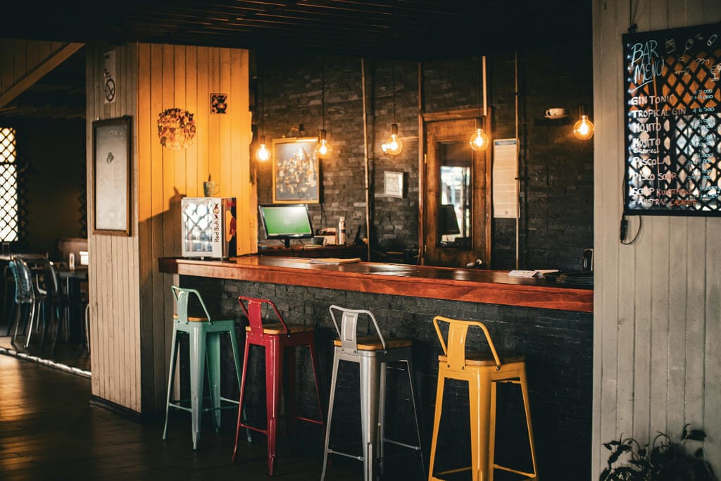 Tunnelmallinen baaritiski, jonka edessä on värikkäitä korkeita jakkaroita ja seinällä valaistu juomalista.