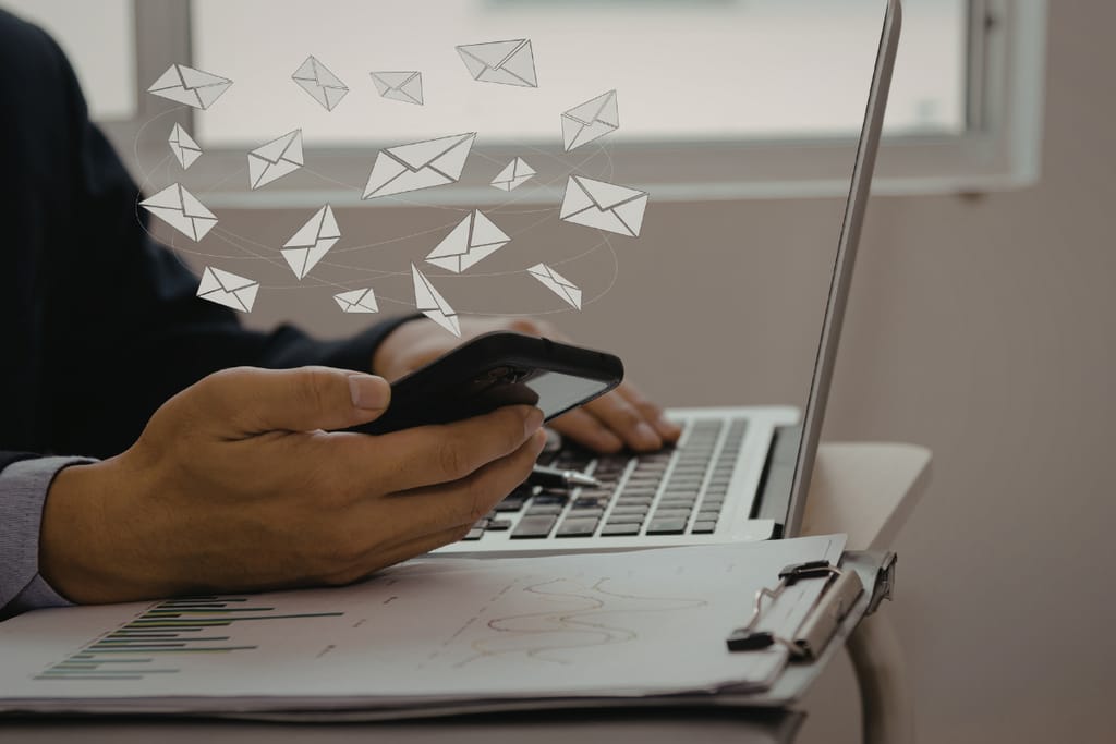 Henkilö käyttää kannettavaa tietokonetta ja älypuhelinta, kuvan päällä leijuu sähköposteja kuvaavia kirjekuvakkeita.