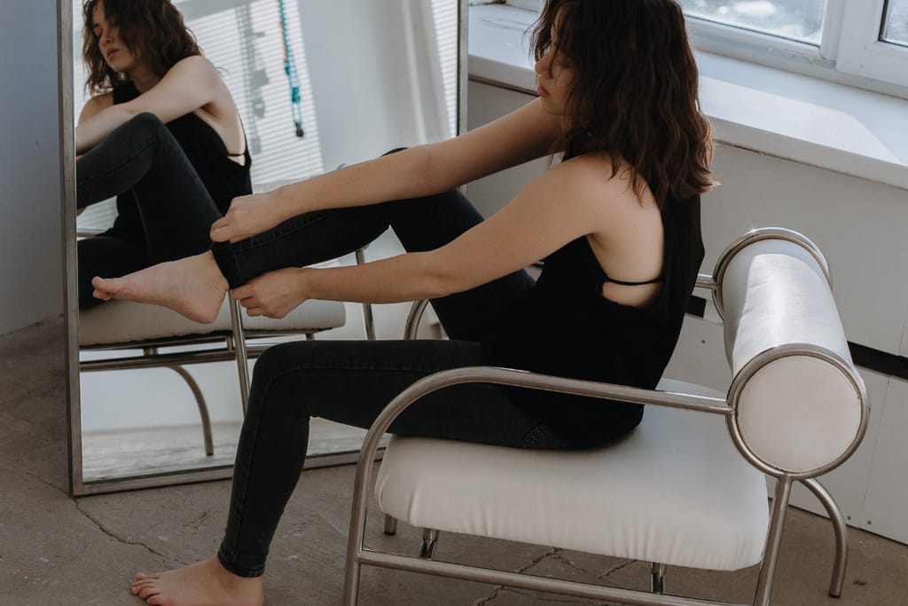Henkilö istuu tuolilla ja vetää tiukkoja farkkuja jalkaan peilin edessä.
