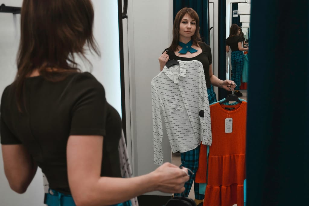 Henkilö katsoo peiliin sovituskopissa ja pitelee kahta eri vaatetta käsissään.