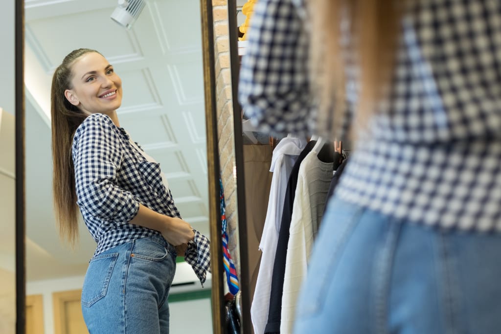 Hymyilevä henkilö sovittaa farkkuja peilin edessä vaatekaupassa.
