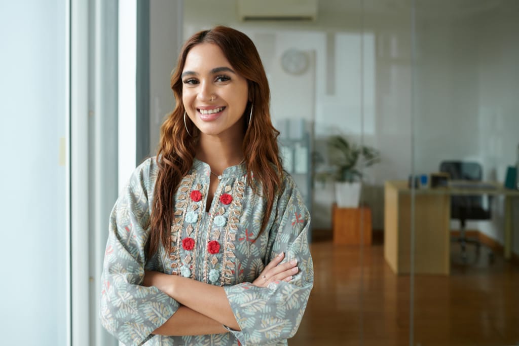 Nainen seisoo sisätiloissa toimistoympäristössä kädet ristissä ja katsoo kameraan hymyillen