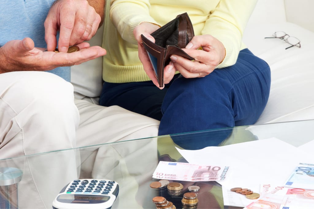 Kaksi aikuista istuu sohvalla ja laskee kolikoita, toinen pitää kädessään tyhjää lompakkoa; pöydällä on euroseteleitä, kolikoita ja laskin.