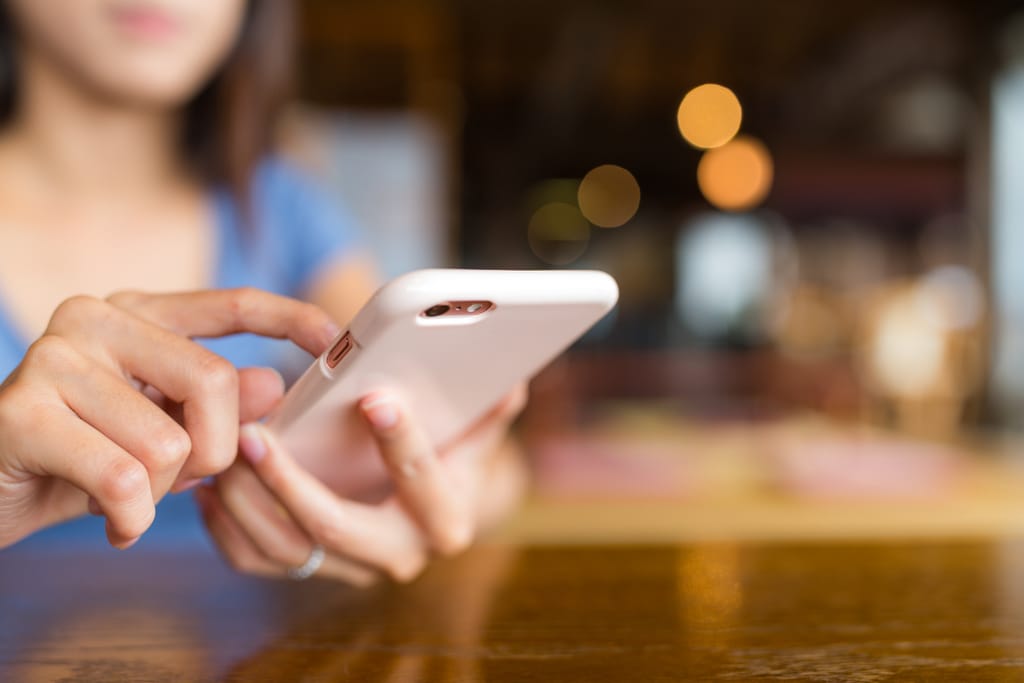 Henkilö käyttää älypuhelinta pöydän ääressä kahvilassa, taustalla epätarkka sisätila ja valoja.