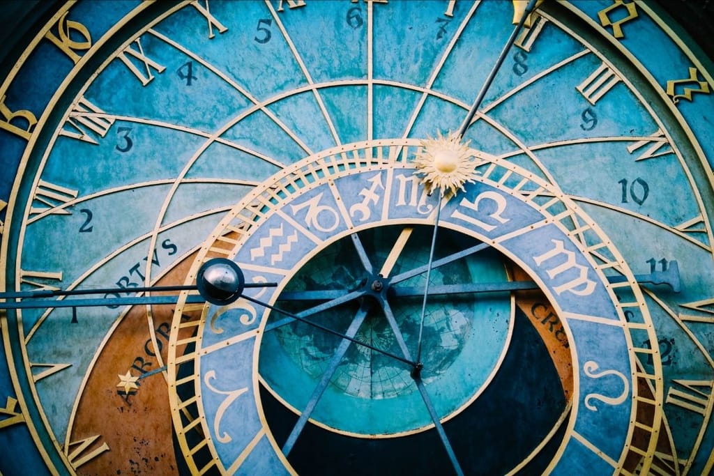 Koristeellinen astronominen kello, jossa astrologisia symboleja ja roomalaisia numeroita turkoosilla taustalla.