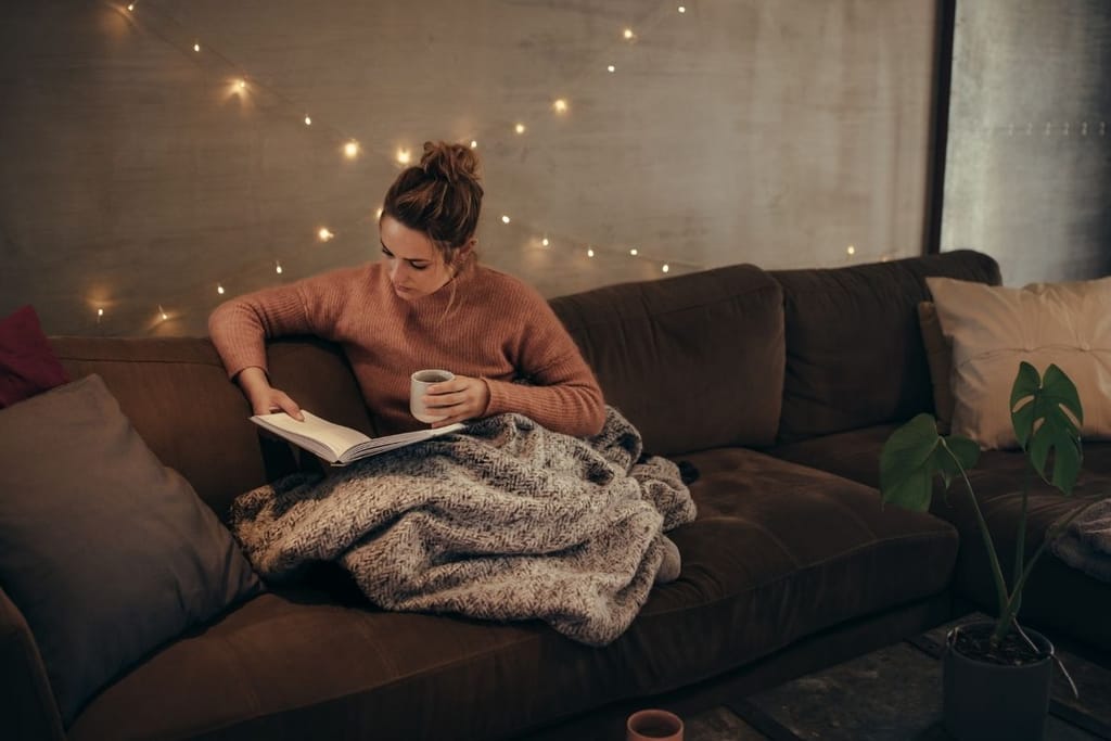 nainen lukee kirjaa yksin kotona sohvalla viltin alla