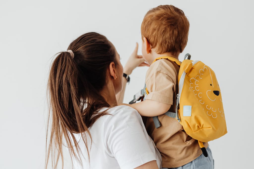 Äiti pitää pientä lasta sylissään, lapsella on selässä keltainen reppu.