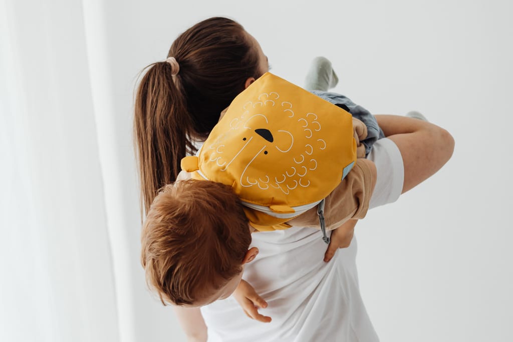 Äiti kantaa pientä lasta, jolla on selässä keltainen leijonakuvioinen reppu.