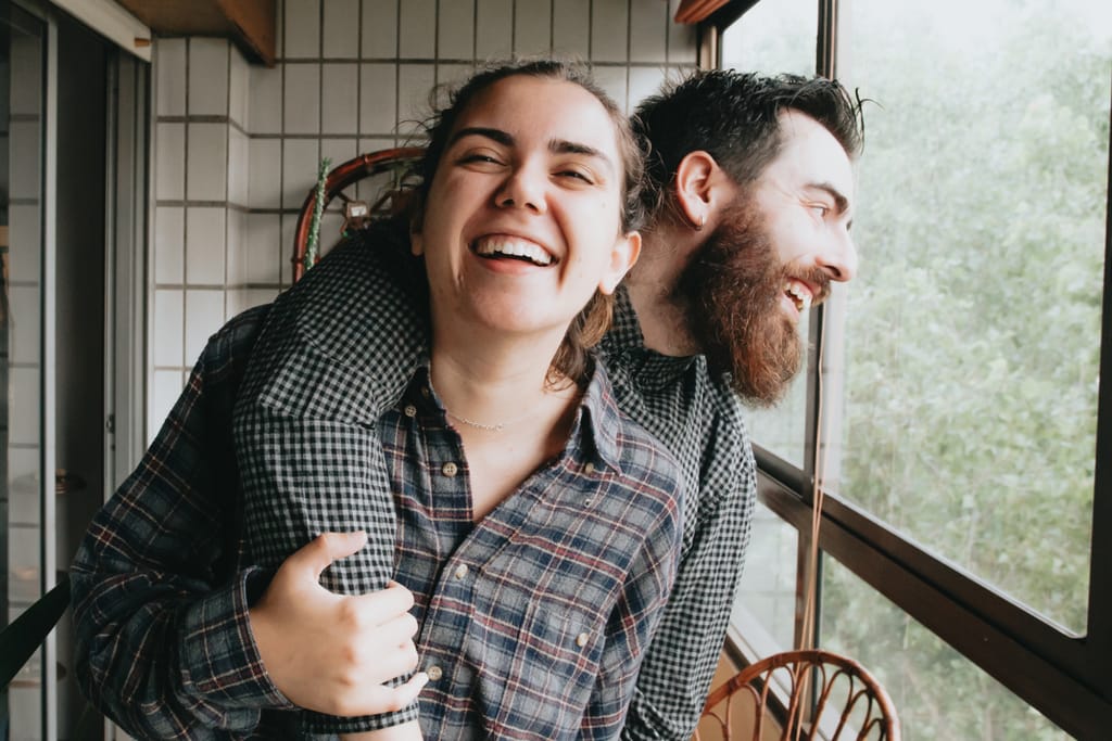 Pariskunta nauraa yhdessä ikkunan äärellä, iloinen ja läheinen tunnelma.