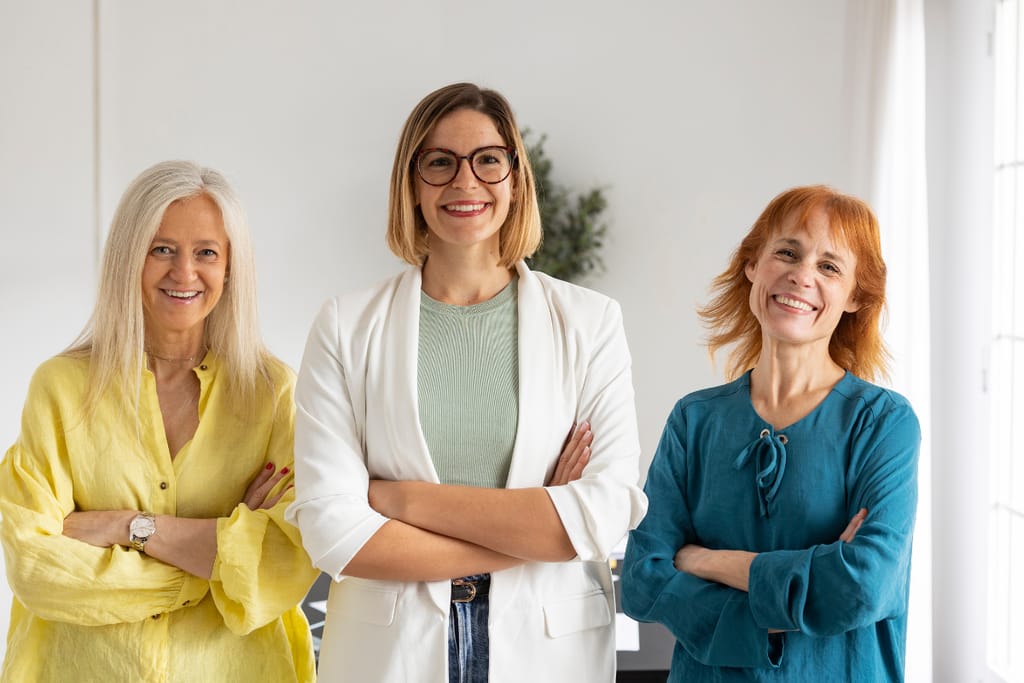 Kolme eri-ikäistä naista seisoo vierekkäin kädet ristissä ja hymyilee kameralle.