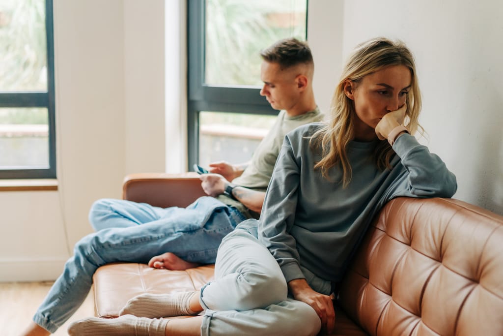 Pariskunta istuu sohvalla etäällä toisistaan, nainen näyttää mietteliäältä ja mies katsoo puhelintaan.