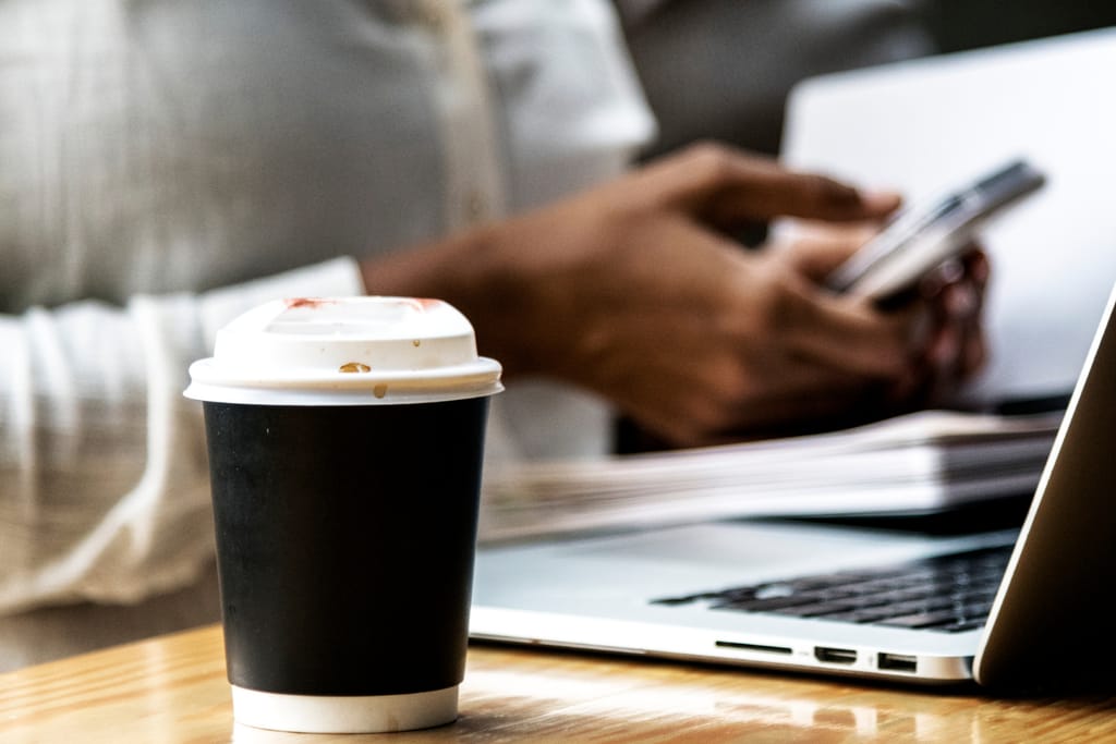 Kertakäyttöinen kahvikuppi pöydällä läppärin vieressä, henkilö käyttää puhelinta taustalla.