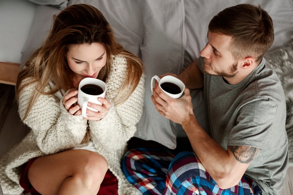 Pariskunta istuu sängyllä ja juo kahvia yhdessä.