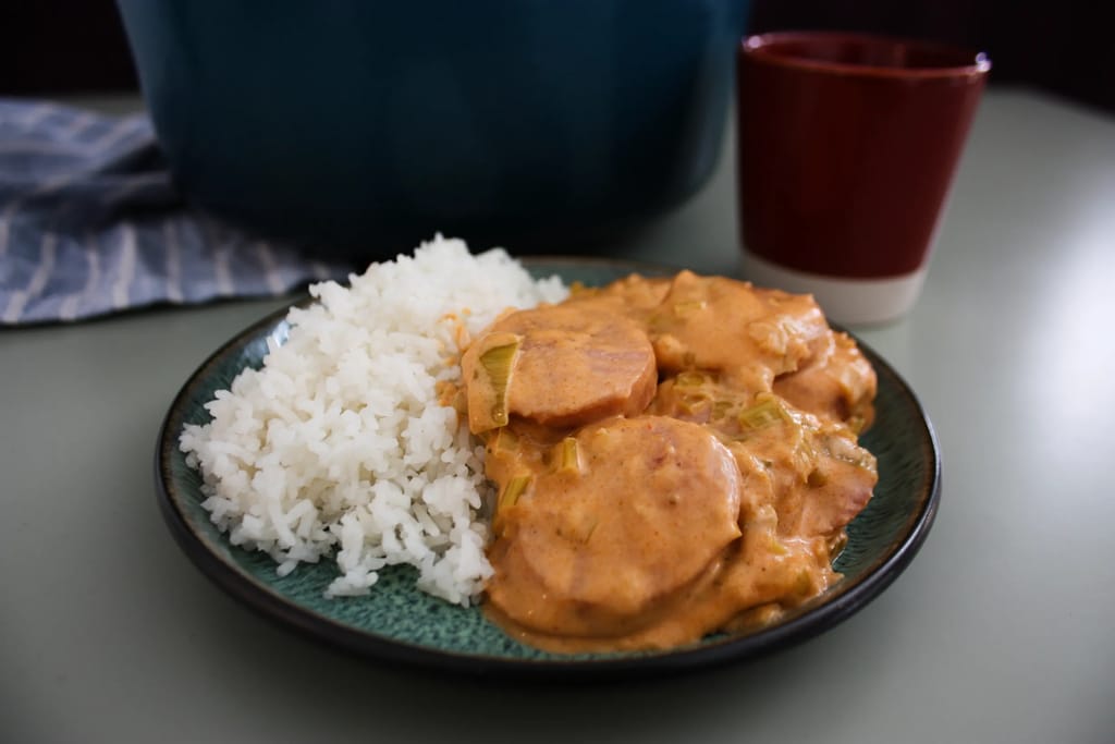 Lautasella riisiä ja kermainen makkarapata; taustalla kattila ja juomalasi pöydällä.