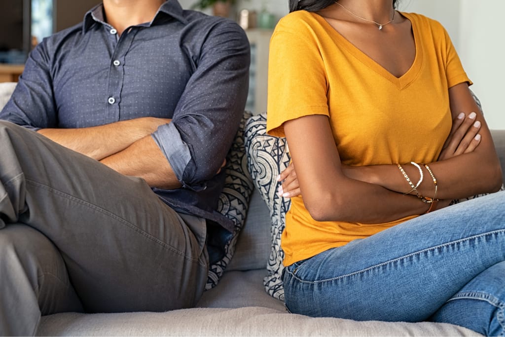 Pariskunta istuu sohvalla selät hieman toisiinsa päin ja kädet ristissä riidan jälkeen.