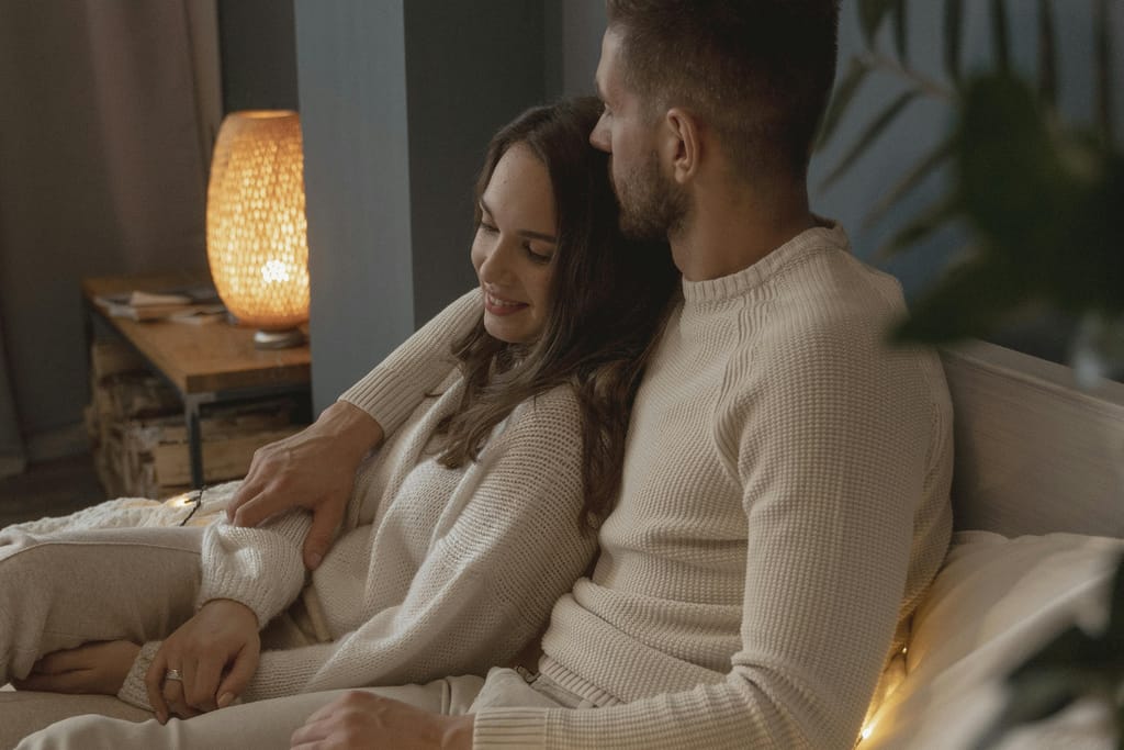 Pariskunta istuu lähekkäin sohvalla ja halailee kodikkaassa valaistuksessa.