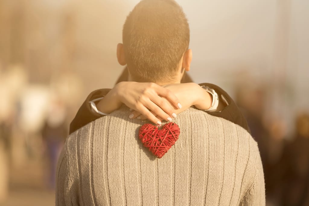 Rakastunut pari kuvattuna miehen takaa, nainen pitää käsissään sydänkoristetta.