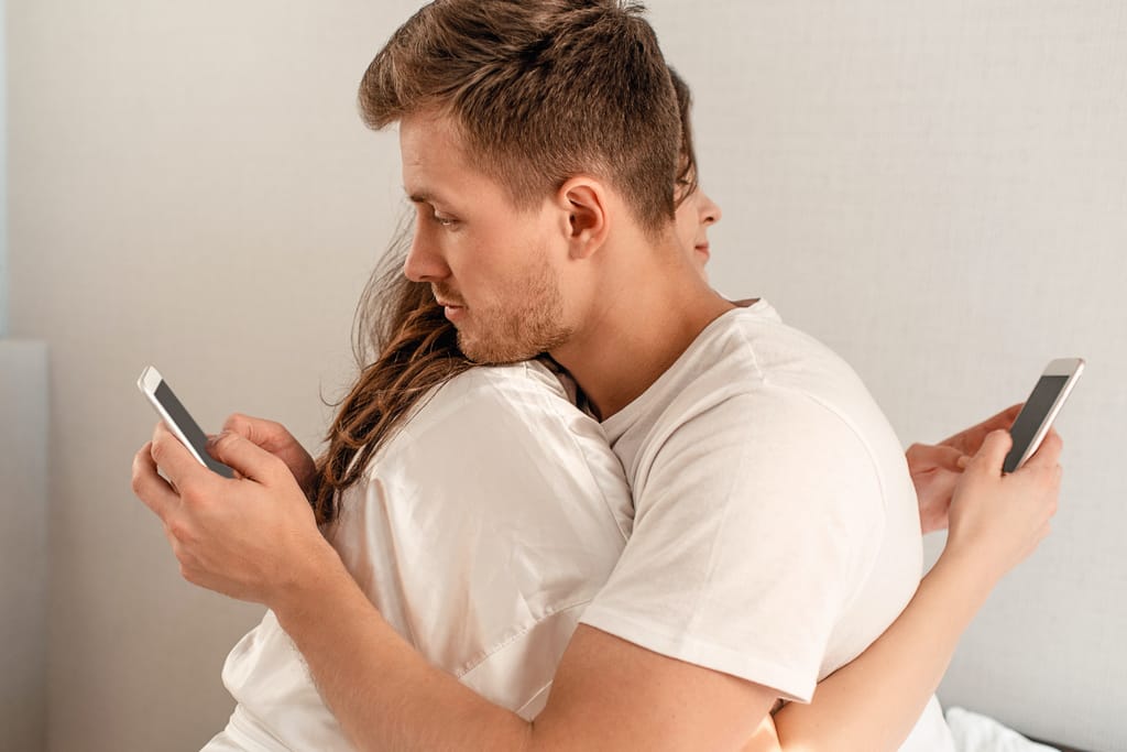 Pariskunta istuu selät vastakkain sängyllä ja katsoo puhelimiaan, etäinen tunnelma.