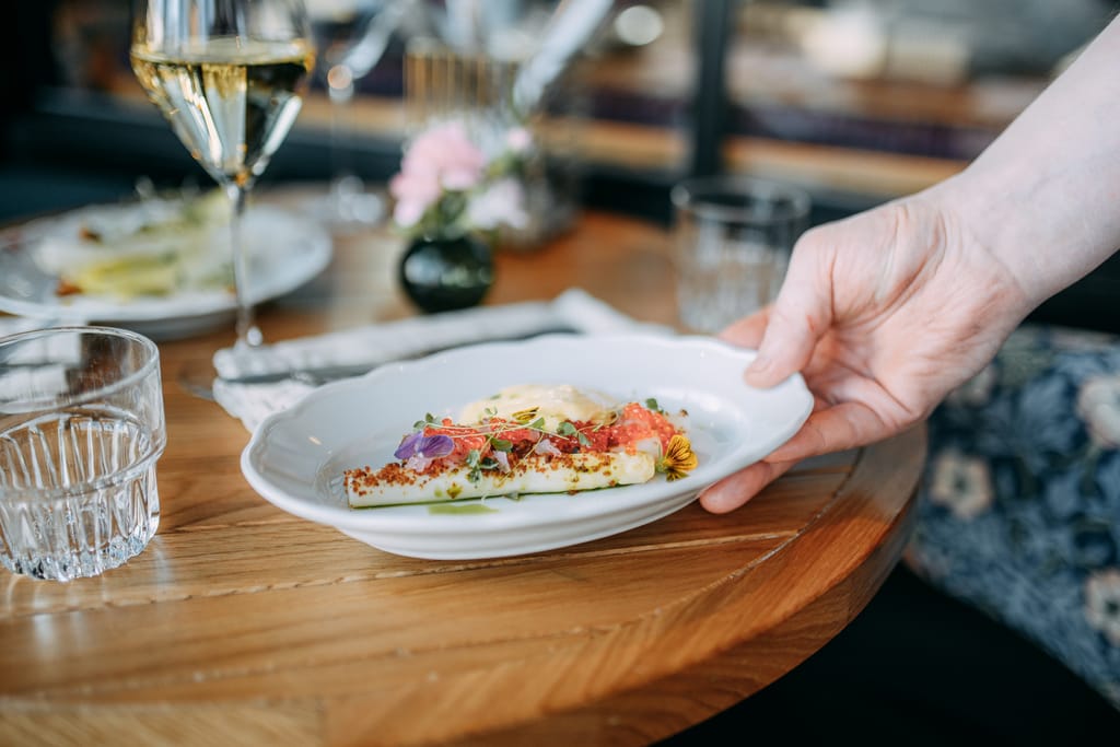 Tarjoilija asettaa ravintolassa pöytään annoksen, jossa on koristeellinen esillepano ja viinilasit taustalla.