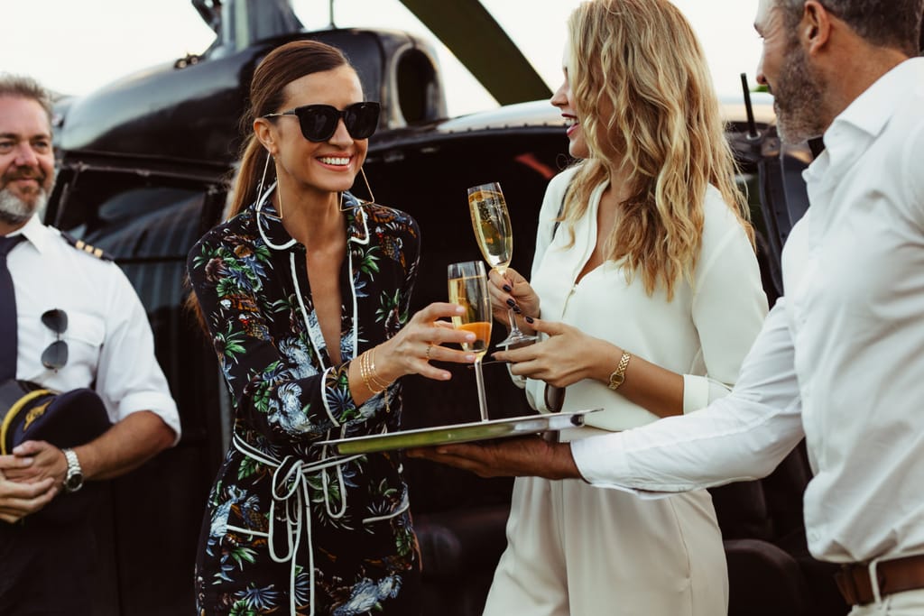 Nainen ottaa tarjottimelta lasillisen samppanjaa. Taustalla helikopteri.