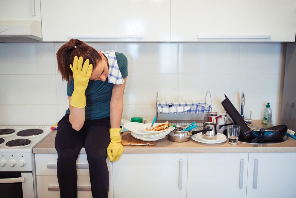 Väsynyt nainen istuu keittiötasolla tiskien keskellä siivoushanskat kädessä.
