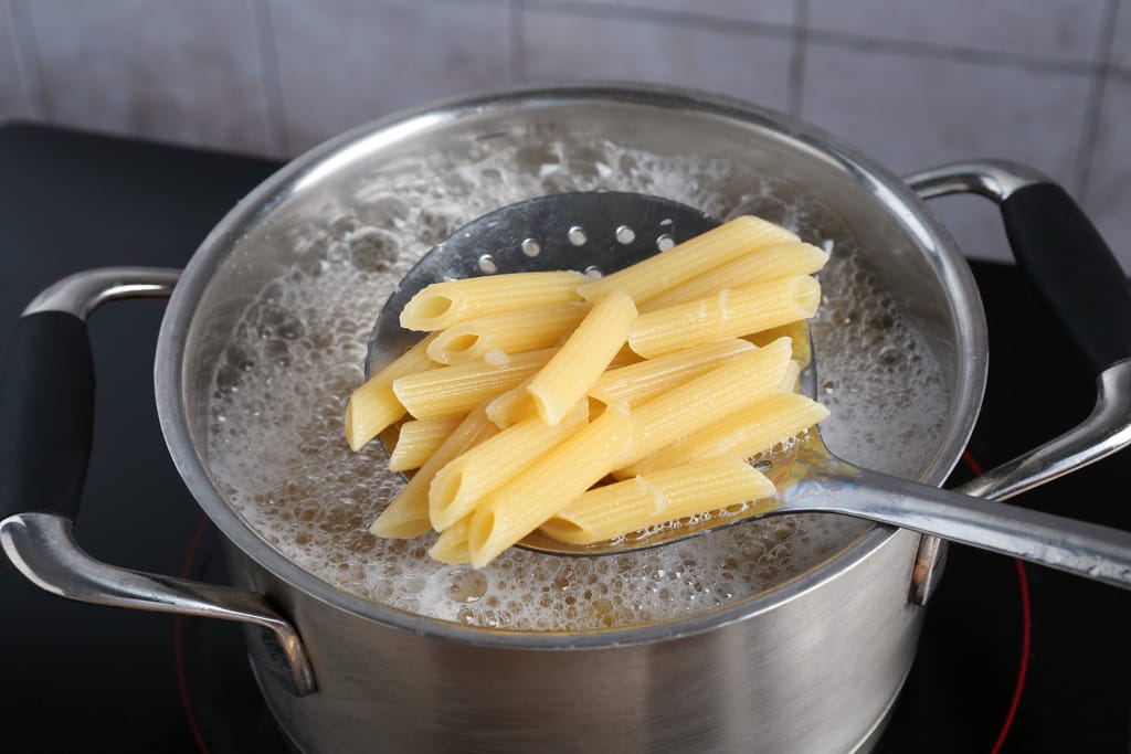 Kattila, jossa kiehuvaa vettä. Nostetaan pastoja lusikalla kiehuvasta vedestä.