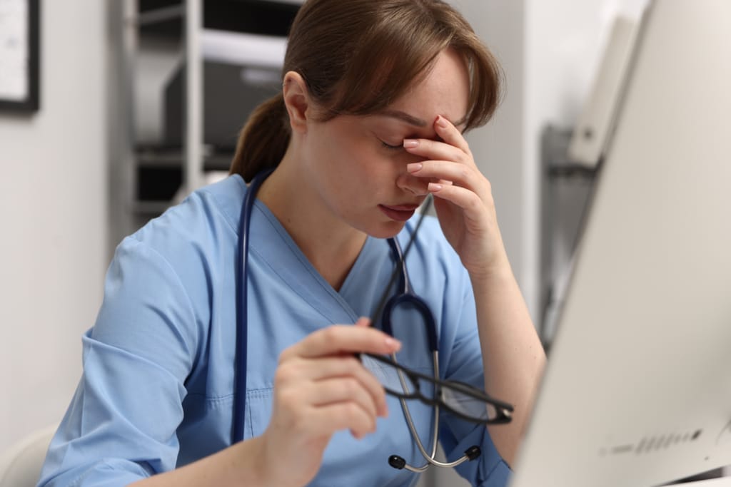 Terveydenhuollon työntekijä istuu tietokoneen ääressä väsyneenä ja pitelee päätään kädessään.