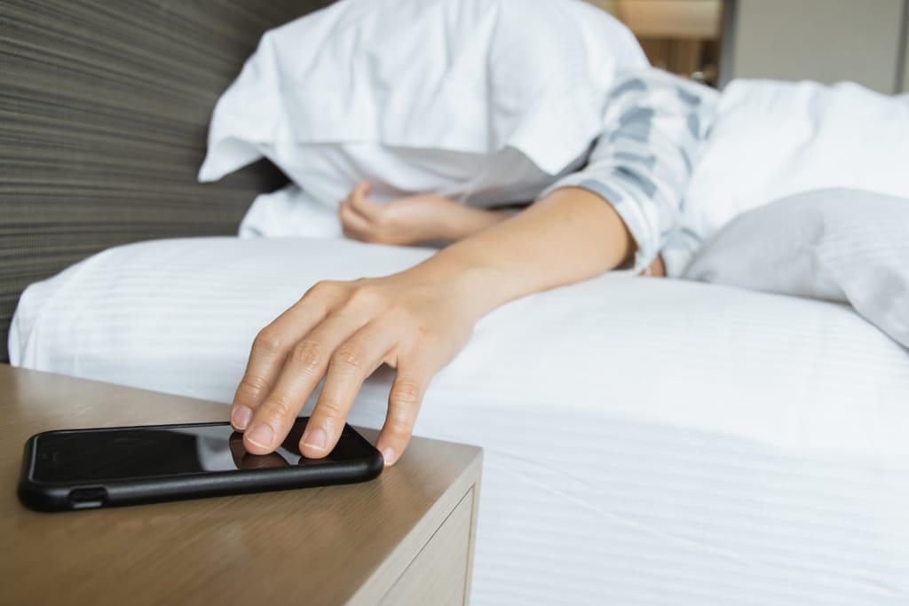 Väsynyt henkilö kurkottaa puhelinta yöpöydältä sängystä käsin.