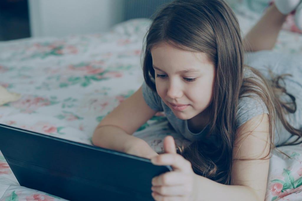 Tyttö makaa sängyllä ja käyttää tablettia keskittyneenä.