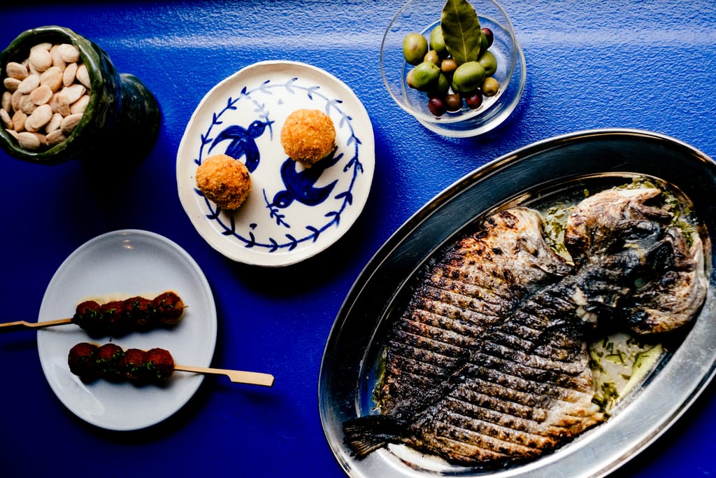 Ylhäältä kuvattu ravintola-annos: kokonaisena grillattu kala hopeavadilla, vieressä pieniä tapas-annoksia – friteerattuja palleroita, lihavartaita, oliiveja ja suolapähkinöitä – sinisellä pöydällä.