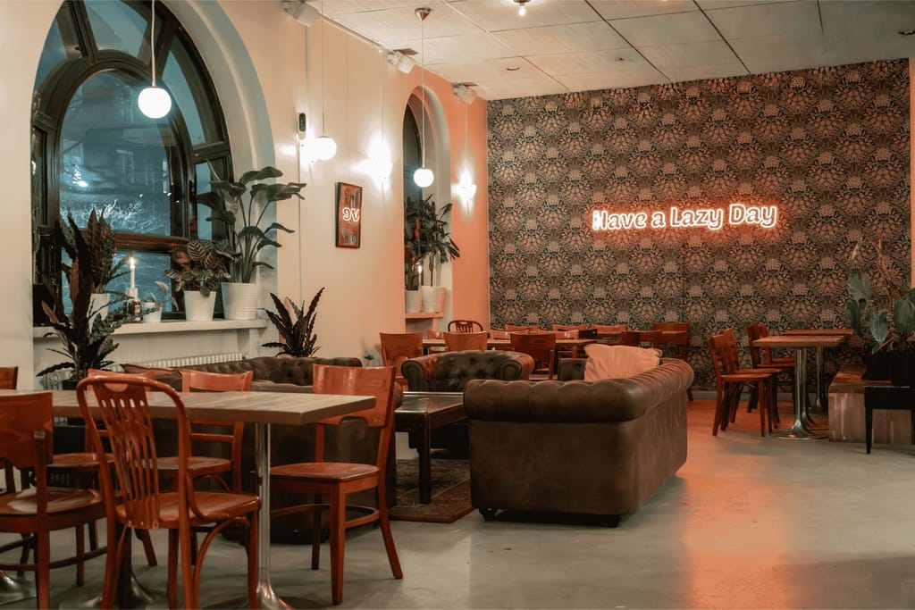 Tyylikäs ja rauhallinen kahvila-ravintolan sisätila, jossa puisia pöytiä ja tuoleja, nahkasohvia, viherkasveja sekä seinällä neonvalo, jossa lukee ”Have a Lazy Day”.