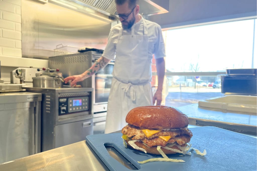 Etualalla kanaburgeri, taustalla kokki valmistaa ruokaa.