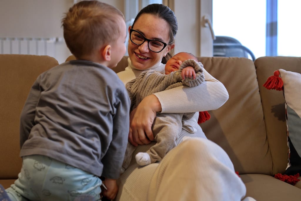 Äiti pitää sylissään vauvaa, isoveli katsoo vierestä.