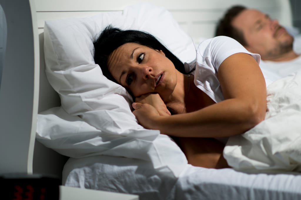 Nainen makaa hereillä sängyssä ja katsoo sivulle, mies nukkuu taustalla – etäinen tunnelma parisuhteessa.