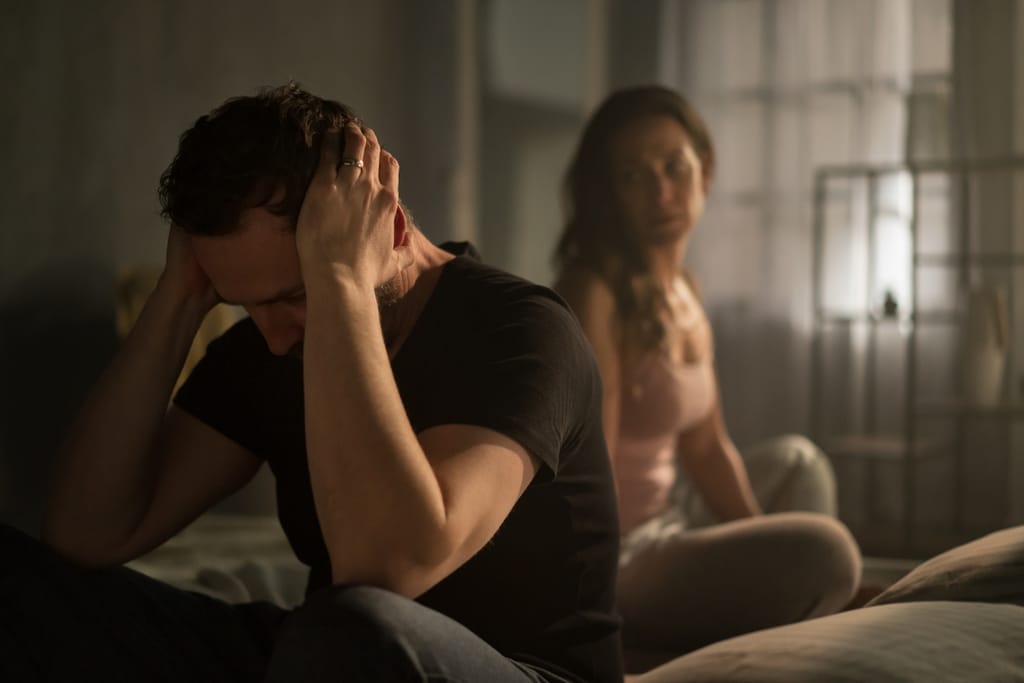 Mies istuu sängyn reunalla pää käsissään, nainen istuu taustalla vakavana – riita tai jännittynyt tilanne parisuhteessa.