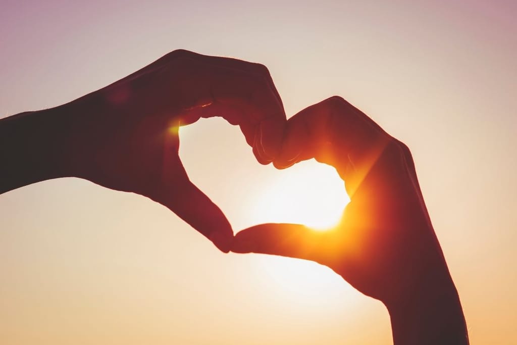 Käsistä muodostettu sydänkuvio auringonlaskua vasten – kaipuu, rakkaus ja yhteys ihmissuhteissa.